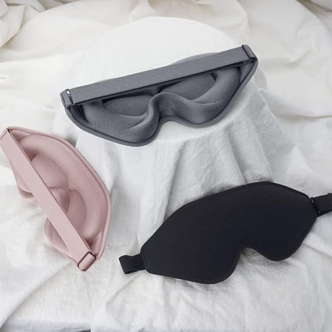 Three eye covers: one black, one blue, and one pink, laid out on a white bed.